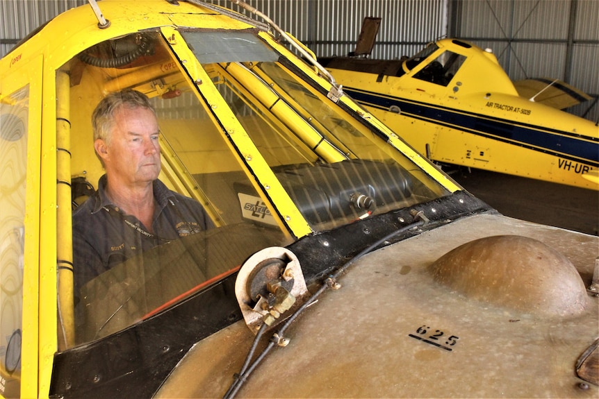 He sits in the cockpit of his yellow plane, which is in a hangar, with another yellow plane in the background. 