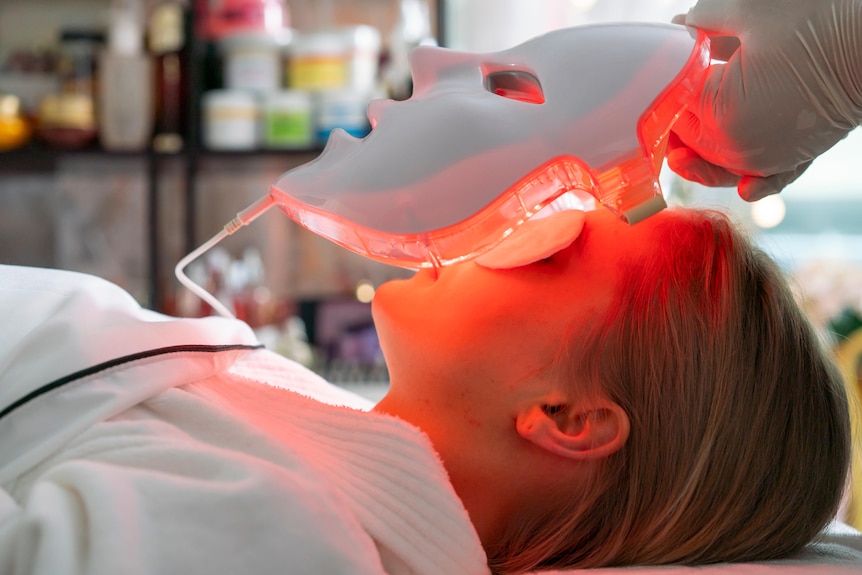 A woman lies on a bed as a red light LED mask is placed on her face.