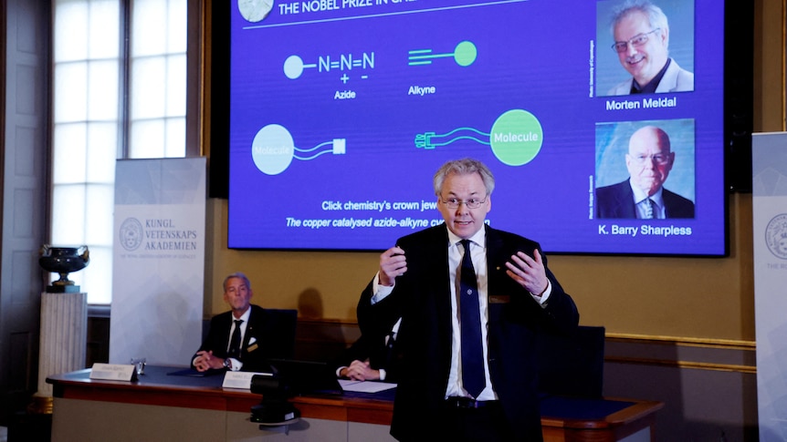 A middle-aged white man in a suit gestures excitedly in front of a screen displaying a chemistry diagram.