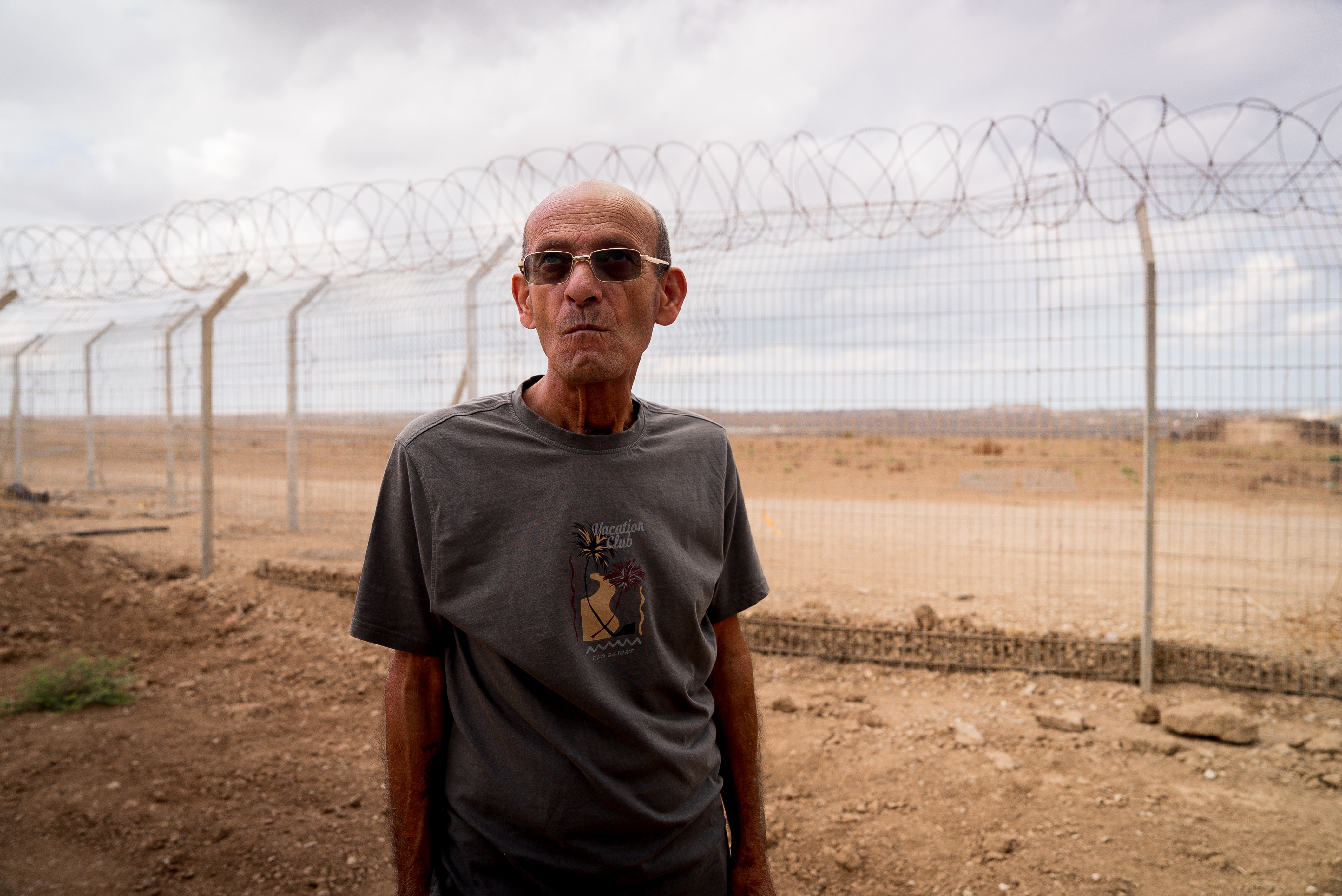 An elderly man wearing sunglasses stands in front of a big wire fence that cuts across a dusty expanse.