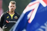 Aaron Finch stands with his hands behind his back in front of an Australian flag