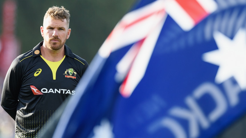 Aaron Finch stands with his hands behind his back in front of an Australian flag