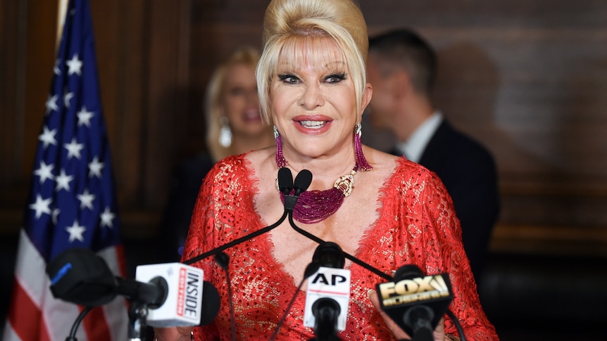 An older, blonde woman with beehive hairdo speaks to media. She's wearing a low-cut red dress, a large necklace and earrings 