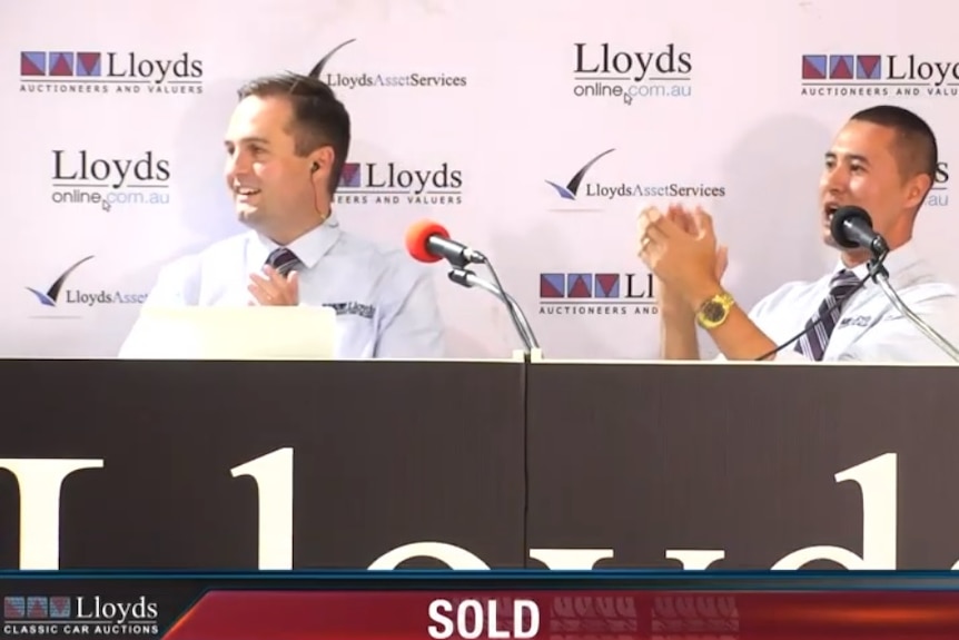 Two men, sitting behind a desk, clapping, in front of a banner that reads 'Lloyds Auctioneers and Valuers'