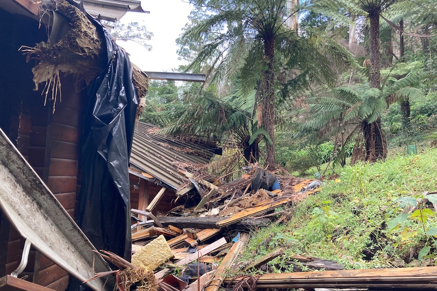 Black plastic, splinters of wooden beams and other building debris is strewn beside a home nestled on a rainforested hill.