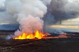 Lava erupts above dark lava fields, sending large plumes of smoke into the atmosphere