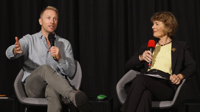 Two people seated on stage with microphones against a dark curtain backdrop.
