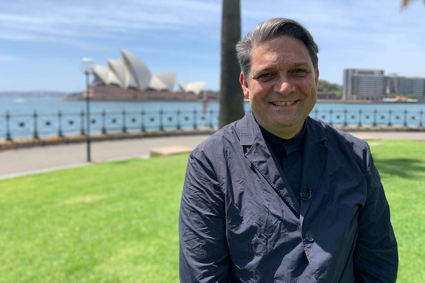 Wesley Enoch stands smiling in a park with the Sydney Opera House and harbour in the background.