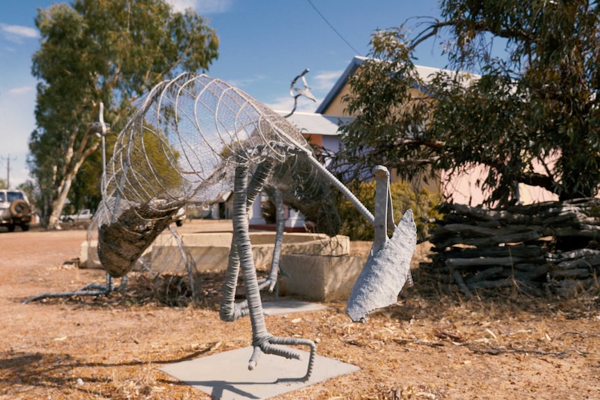 Metal emu sculptures outdoors on gravel, a colourful building, a tree and a four wheel drive in the background