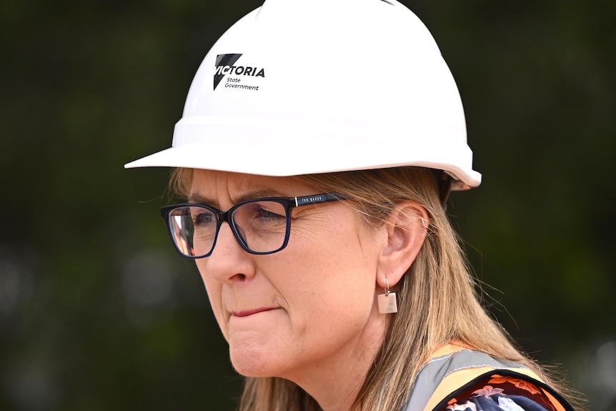 Jacinta Allan appears thoughtful, wearing a white hard-hat and high-vis vest at an outdoor press conference.