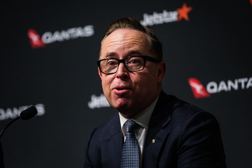 Alan Joyce wears glasses and a suit and sits in front of a black background which says Qantas on it.
