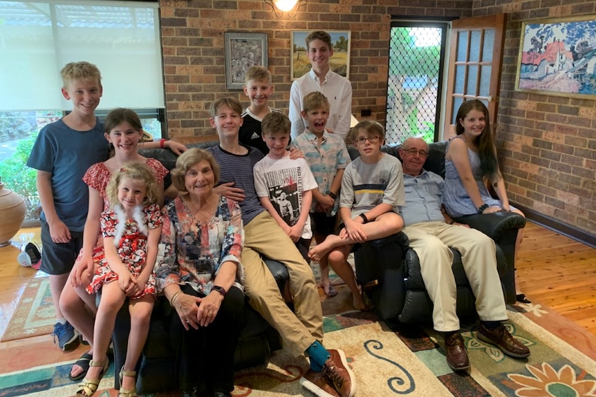 Family of 10 children and two grandparents sit black couch in room with brick walls and wooden floorboards