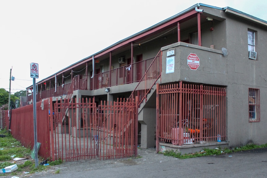 A three-story run-down apartment building is surrounded by a security gate and trash.