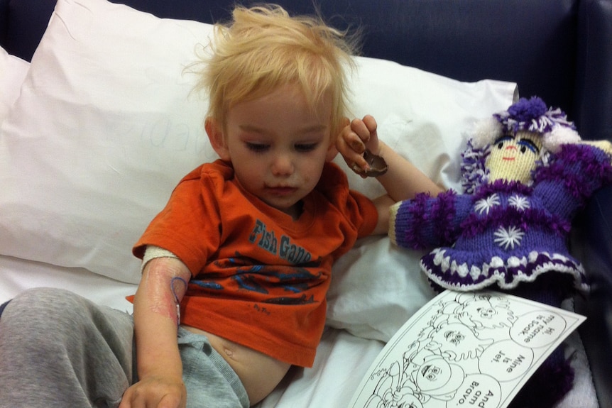 Young boy with blonde hair lies on bed reading a book with purple toy beside him.