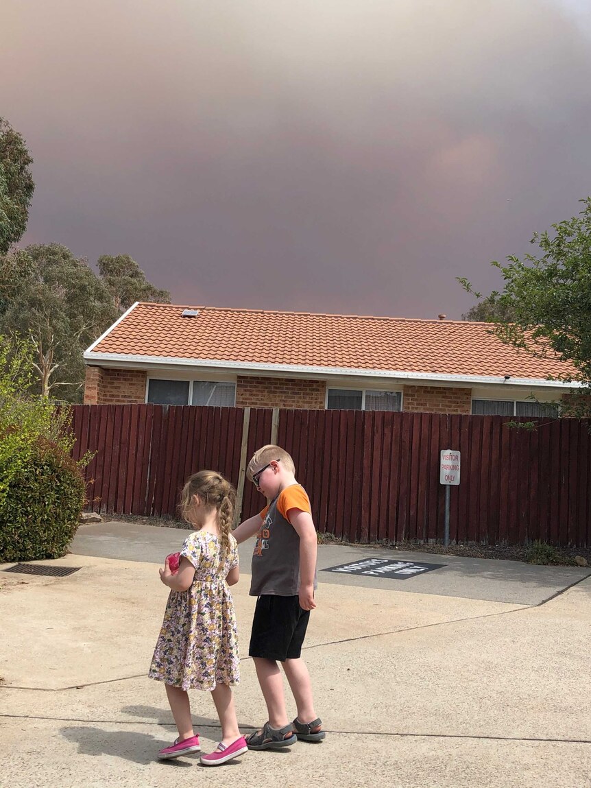 Charlotte and Jeremy walk down their driveway, a huge plume of smoke and signs of flames in the background behind some houses.