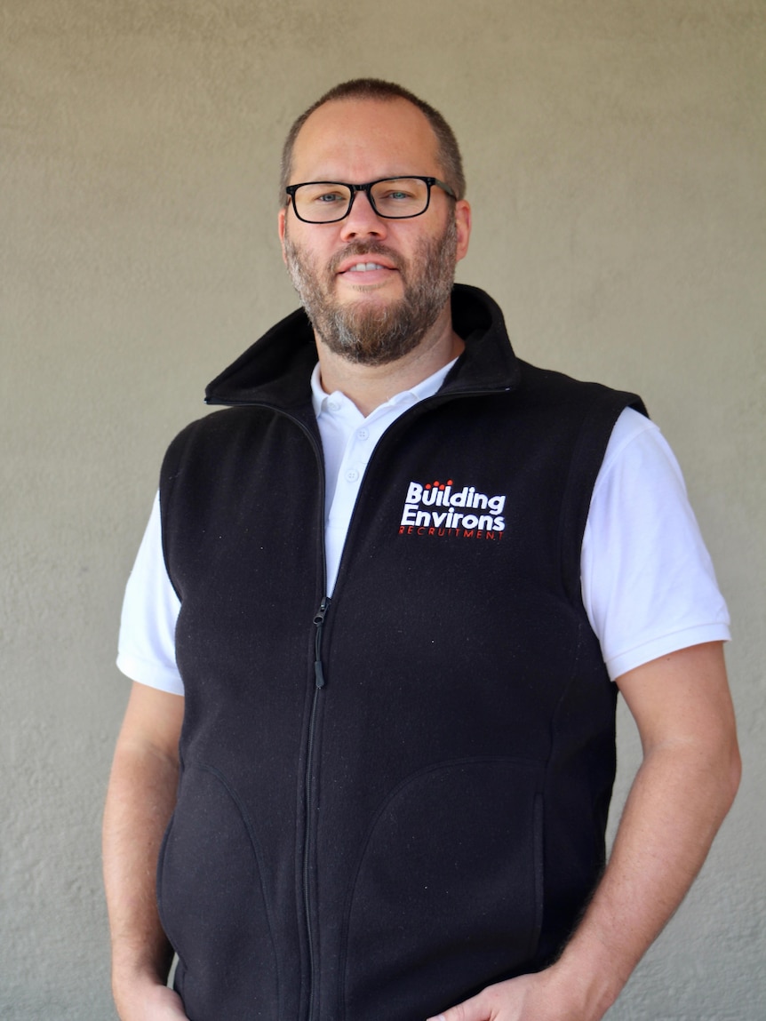 Portrait of a middle-aged man in a white shirt and a warm vest. He has glasses on and a grey short beard.