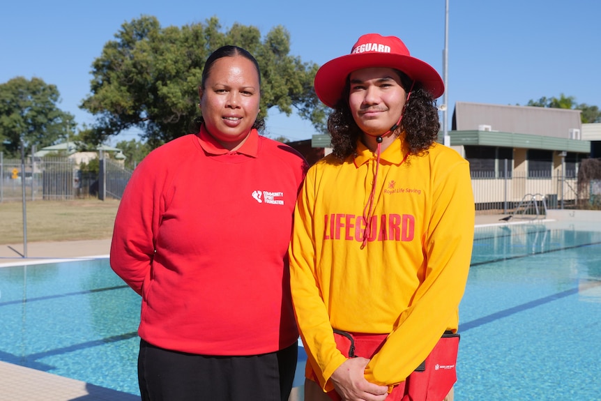 A woman and man stand in front of a swimming pool. The woman wears a red shirt and the man is wearing a yellow lifeguard shirt. 