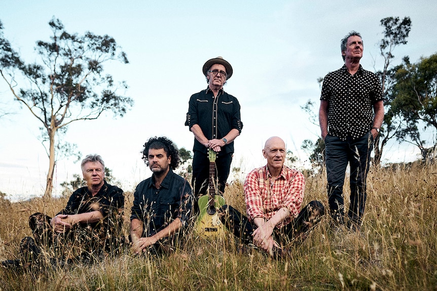 Members of Midnight Oil in a field of long grass. Three members sit and two of them stand.