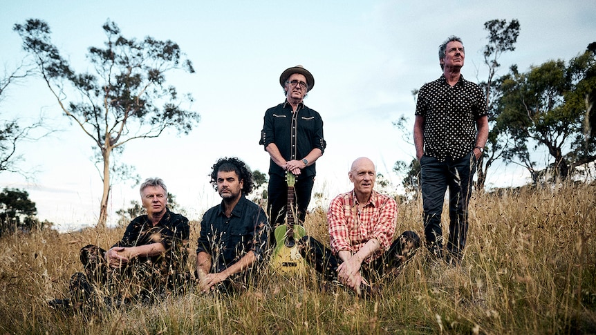 Members of Midnight Oil in a field of long grass. Three members sit and two of them stand.