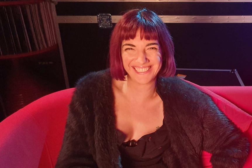 A woman with short brown hair and a fringe smiling and sitting on a red couch.