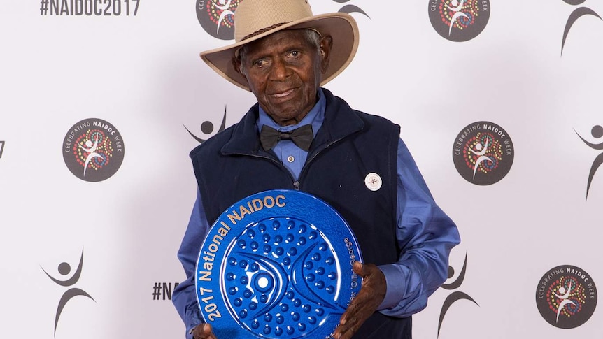 Badimaya elder Alex "Ollie" George stands holding blue award.