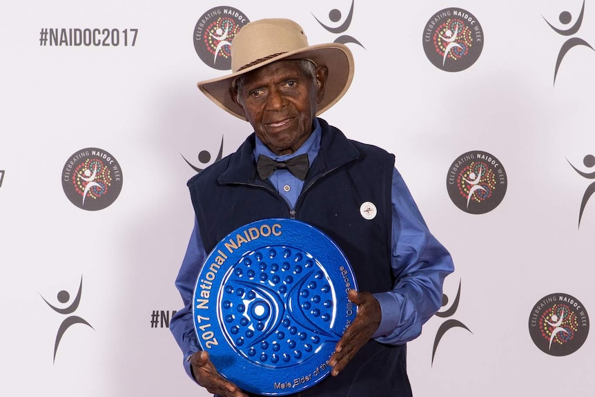 Badimaya elder Alex "Ollie" George stands holding blue award.