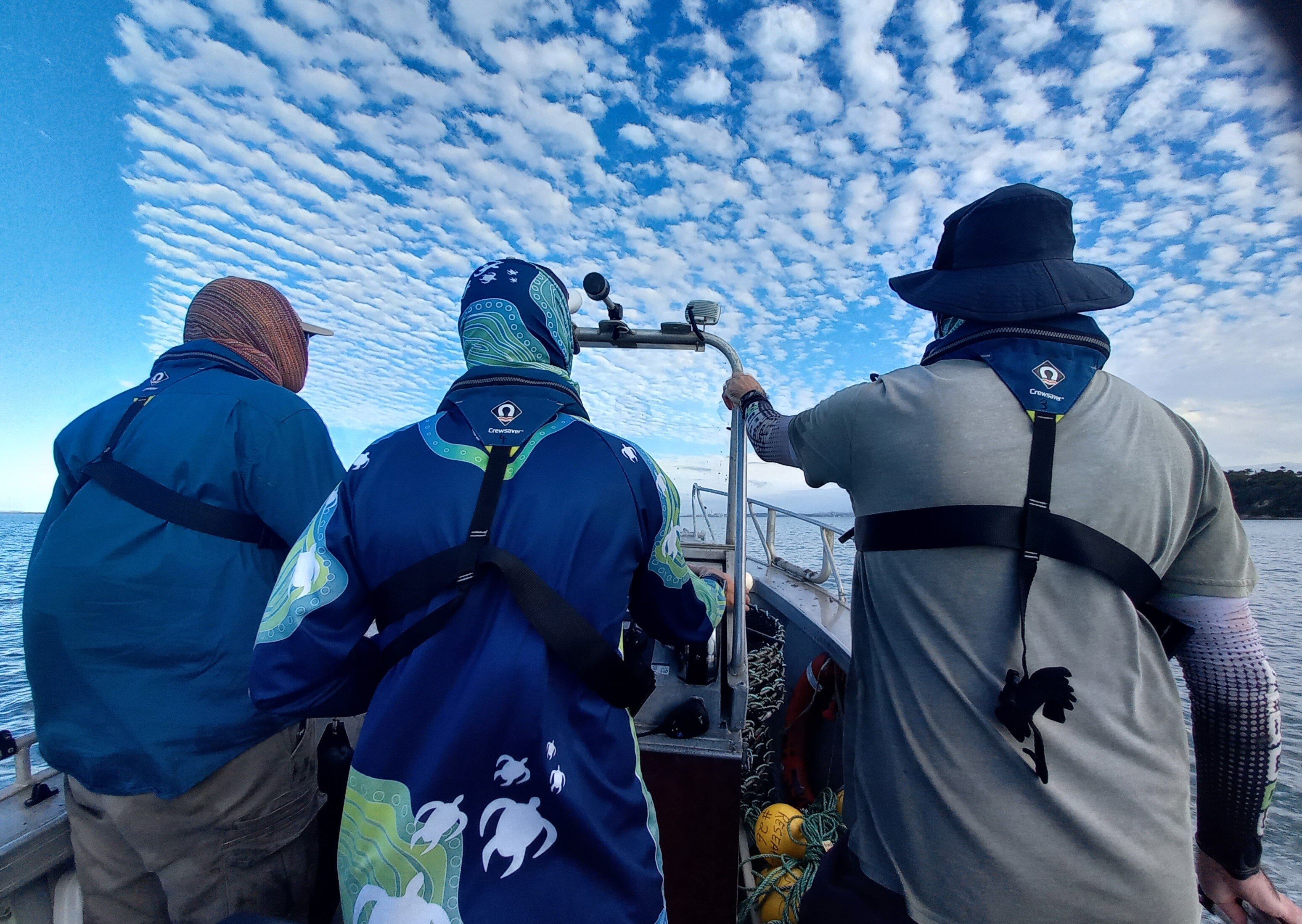 The back of three men in UV protective clothing on a boat with clear blue sky and white clouds. 