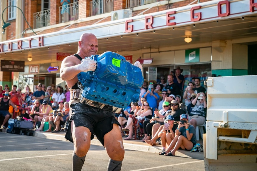 Man lifting car engine in front of a large crowd