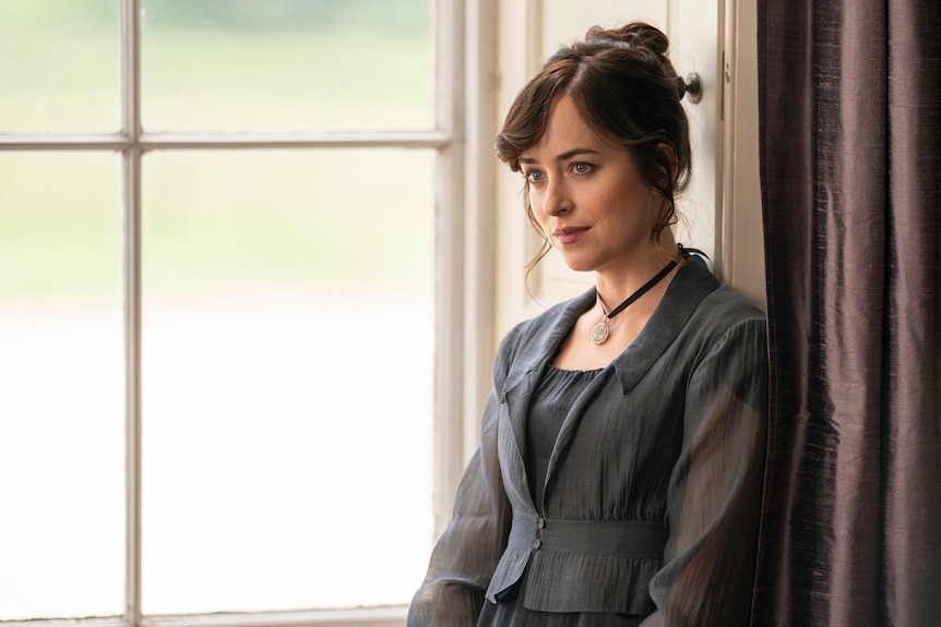 A woman in a gown stands by a window looking thoughtfully at someone in front of her.