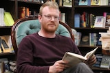 Toby Wools-Cobb sits in a grand old arm chair, surrounded by books, while reader a chunk old title.