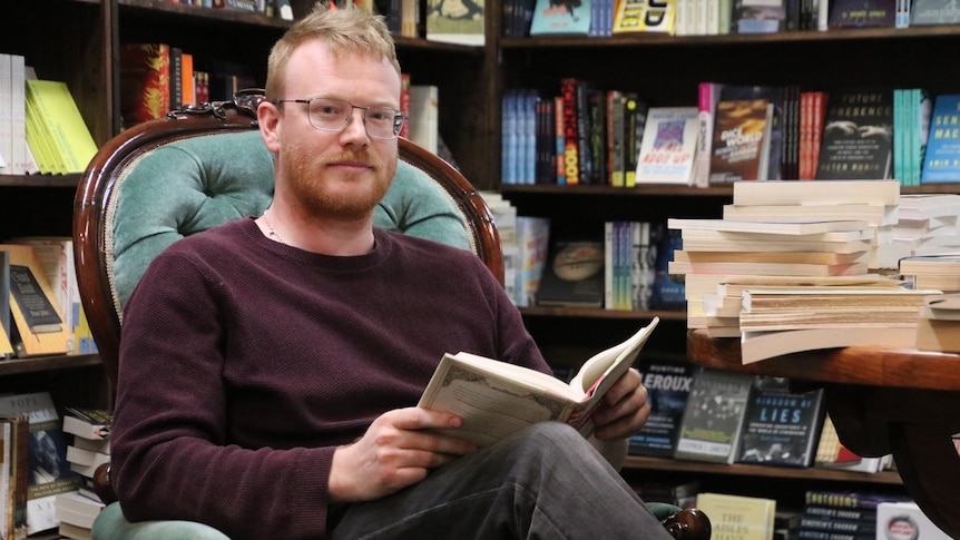 Toby Wools-Cobb sits in a grand old arm chair, surrounded by books, while reader a chunk old title.