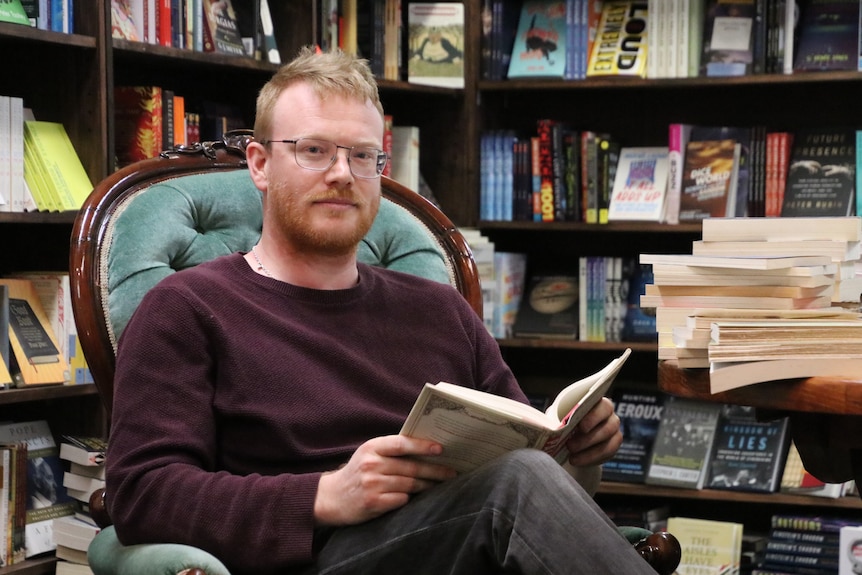 Toby Wools-Cobb sits in a grand old arm chair, surrounded by books, while reader a chunk old title.