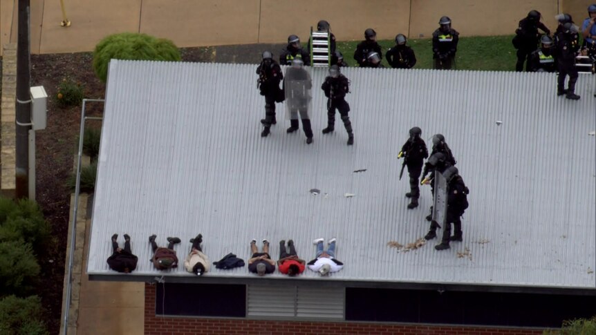 Aerial vision of police in riot gear on top of a roof at Banksia Hill Detention Centre, multiple detainees lying on stomachs.