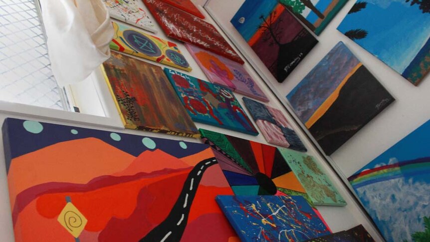 Low angle photo of a wall covered in brightly coloured paintings. Painting called long road home hangs in the left hand corner.