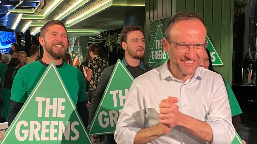 Adam Bandt smiles as he clasps his hands, standing in front of Greens supporters at a party event.