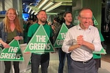 Adam Bandt smiles as he clasps his hands, standing in front of Greens supporters at a party event.