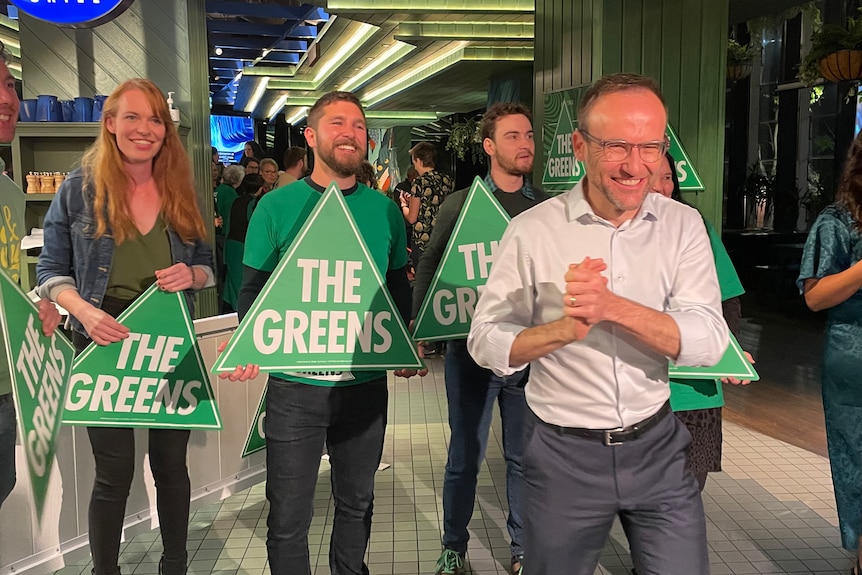 Adam Bandt smiles as he clasps his hands, standing in front of Greens supporters at a party event.