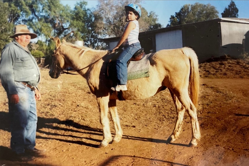Chelsea as a kid, sitting on a white horse, man standing to her left, dirt on the ground.