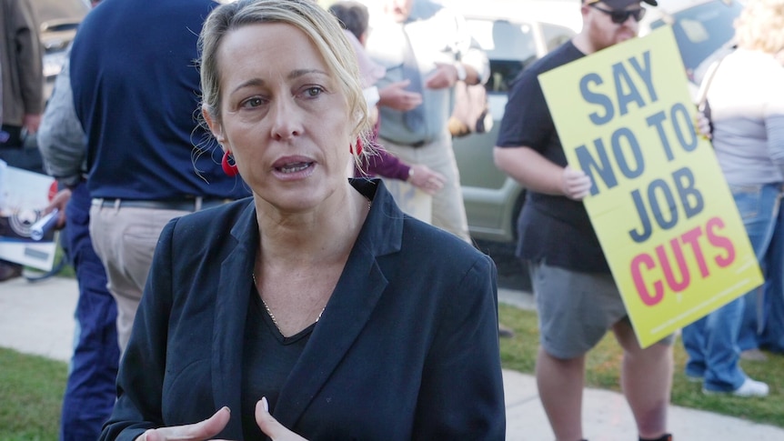 A middle aged blonde woman in a dark blazer looks frustrated as she speaks a protester holds a say no to job cuts sign