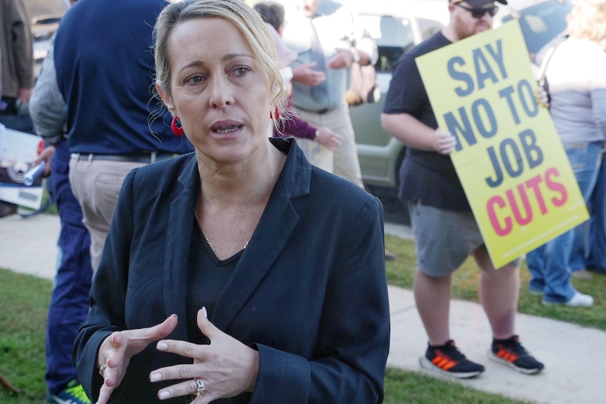 A middle aged blonde woman in a dark blazer looks frustrated as she speaks a protester holds a say no to job cuts sign