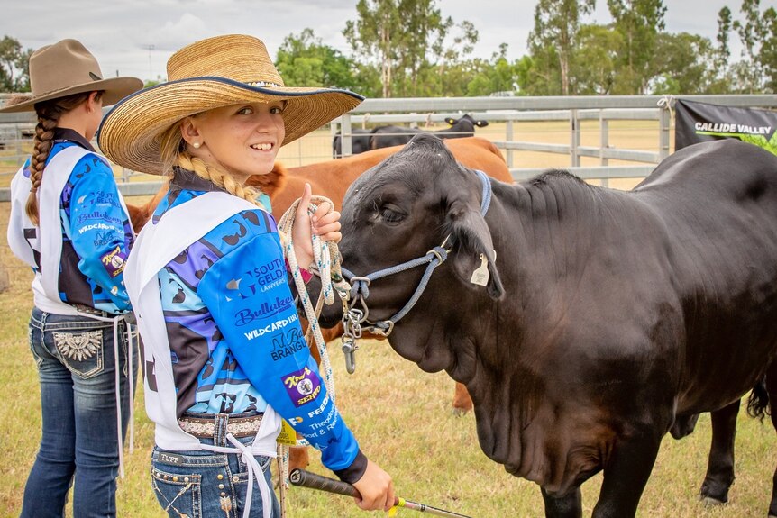 Ella wearing a blue shirt, wide brimmed hat, holding holding rope attached to the cow next to her.
