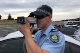 A police officer wearing sunglasses holds up a radar gun to his eye by the side of the road