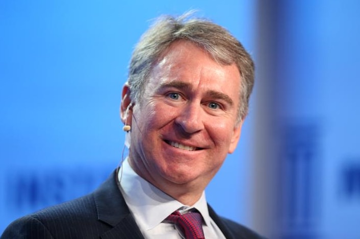 Ken Griffin looks directly to the camera with a smile. A microphone is in his ear and he wears black suit against blue backdrop.