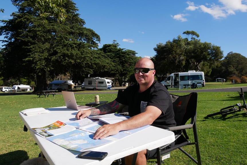 A smiling bald man in a black t-shirt, sunglasses, sits on a folding chair, a table with maps in the trailer park. 