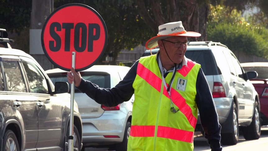 Rodney James in sun hat and fluoro vest holds a stop sign at a school crossing