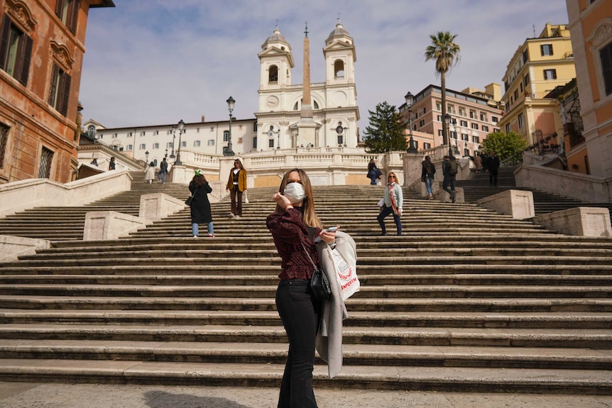 A young woman wearing a mask and carrying a plastic bas stands at the bottom of steps.