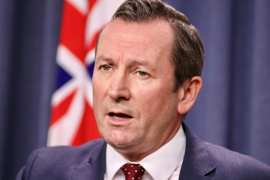 A close up of Mark McGowan wearing a suit, standing in front of a blue background and Australian flag. 