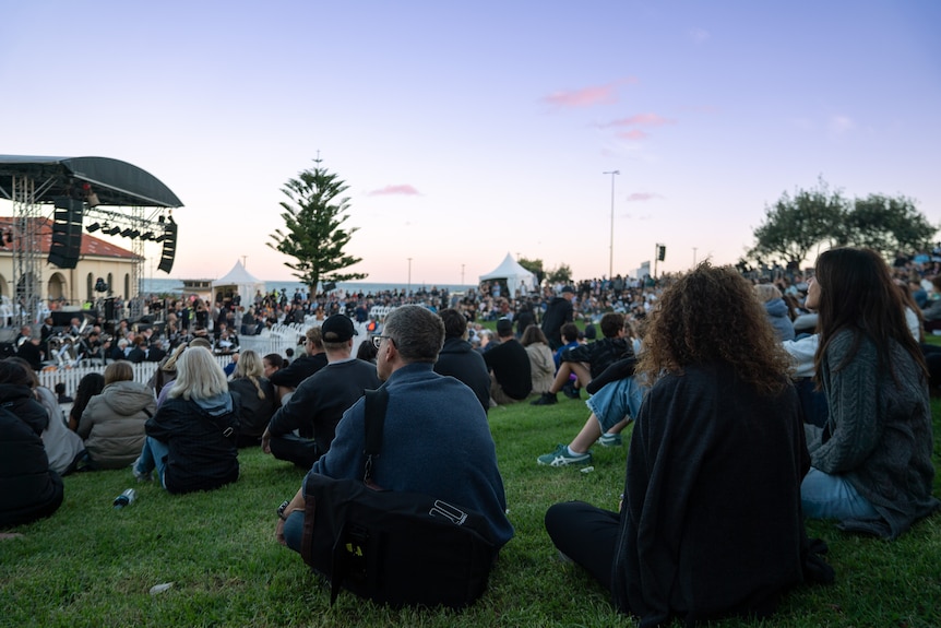 A crowd of people sit on grass in front of a stage, with the beach visible in the background.