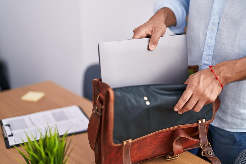 A man standing beside a timber desk puts a silver laptop into a brown leather bag.