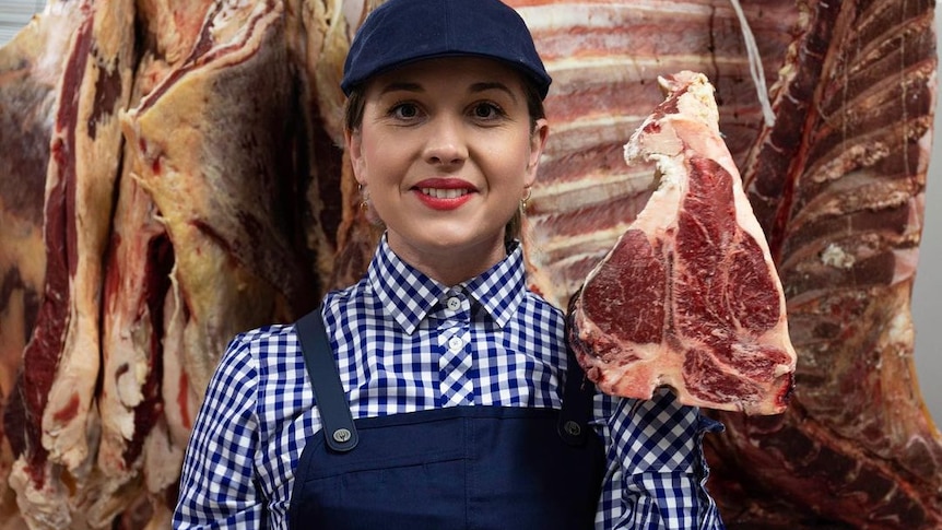 A smiling woman holding a cut of meat, carcass and meat hanging behind her.